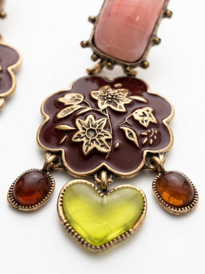 A close-up of an ornate jewelry piece featuring a scalloped dark red enamel flower with gold-tone floral detailing, a large light green heart-shaped stone, and two amber oval stones, with a pink stone at the top.