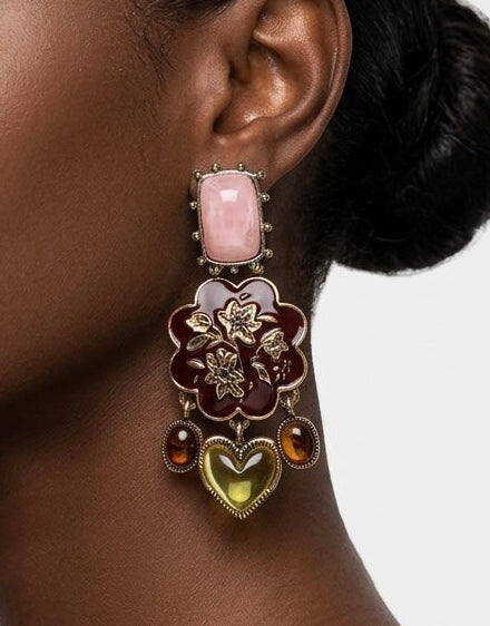 A close-up side profile of a woman wearing a large, ornate statement earring with pink, gold, and brown floral details, against a light gray background.